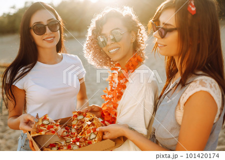 Three young woman spending time together at the beach, having picnic, eating pizza. 101420174