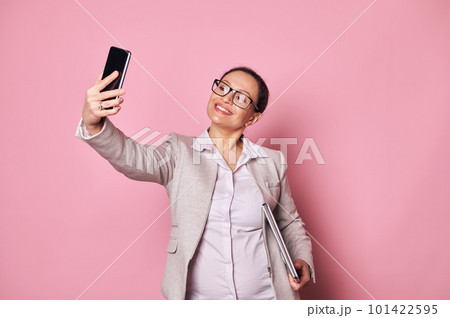 Attractive middle-aged multi-ethnic pregnant woman, business person wearing casual clothes and trendy glasses, carrying laptop and taking selfie on her smartphone on isolated pink background. Ad space Attractive middle-aged multi-ethnic pregnant woman, business person wearing casual clothes and trendy glasses, carrying laptop and taking selfie on her smartphone on isolated pink background. Ad space 101422595