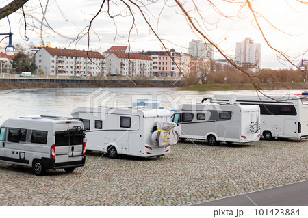 Many white modern campervan recreational motor home vehicles parked in row at camper park site Magdeburg city against Elbe river bridge. Motorhome campground stataion travel destination. RV lifestyle 101423884