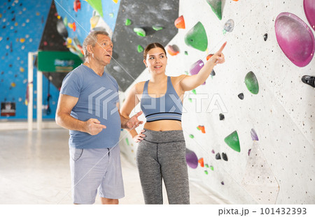 Friendly young female coach of climbing gym instructing aged man 101432393