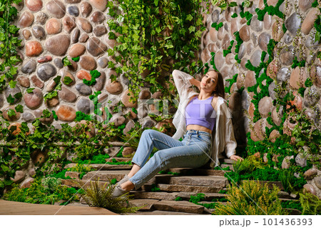 Cute woman sits on stone plate steps with old overgrown stone wall in the park 101436393