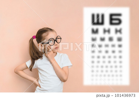 Thoughtful smart girl of elementary school age in round glasses against the background of a blurred sivtsev table for checking visual acuity Thoughtful smart girl of elementary school age in round glasses against the background of a blurred sivtsev table for checking visual acuity 101436973