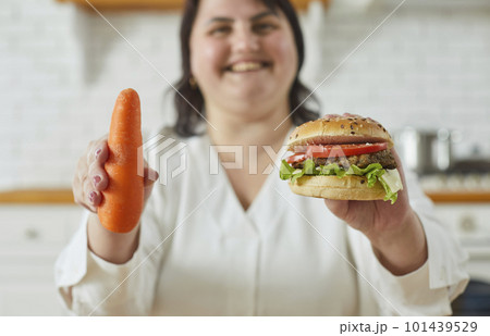 Young overweight plus size fat woman holding a burger in one hand and a carrot in other. 101439529