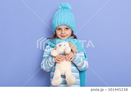 Image of charming little female, child with fluffy puppy in hands, looking directly at camera, dresses casual clothing, scarf and hat, posing isolated over lilac studio background. Childhood concept. 101445238