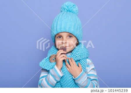 Closeup portrait of sweet cute blue eyed little girl wearing blue scarf, hat and striped sweater, holding food in hands, eating sweets, looking directly at camera. Children and winter concept. 101445239