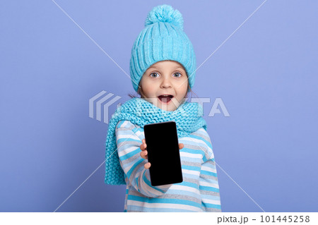 Astonished little girl kid wearing winter hat with pom pom showing blank screen for text space of new popular mobile phone isolated over blue background, child standing with opened mouth and surprised 101445258