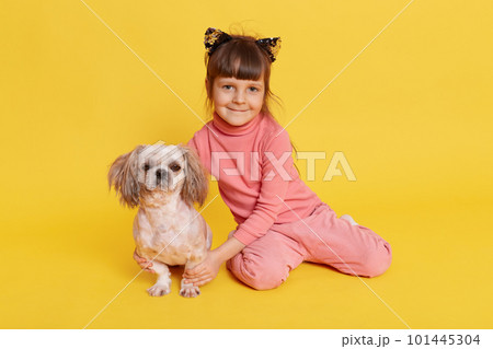 Little dark haired girl with puppy posing indoor, child looking at camera and smiling, hugging her pet, wears casual attire and hair band with cat's ears, isolated over yellow background. Little dark haired girl with puppy posing indoor, child looking at camera and smiling, hugging her pet, wears casual attire and hair band with cat's ears, isolated over yellow background. 101445304