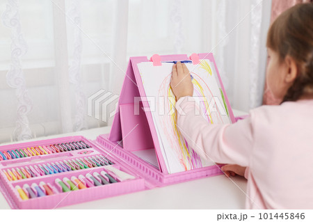 Back view of child with pigtails painting picture on paper from set of art and creativity, many multicolored pencils and wax crayons, kid sitting at table indoor. 101445846