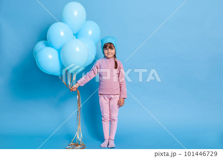Dark haired little girl in cap and pink suit, holds big bunch of helium balloons, being on children's party, looks shyly at camera, full length of female child celebrates birthday. Dark haired little girl in cap and pink suit, holds big bunch of helium balloons, being on children's party, looks shyly at camera, full length of female child celebrates birthday. 101446729