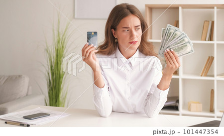 Online payments. Hesitating woman. Money keep. Puzzled lady choosing between credit card and cash dollars sitting work desk in light room interior. 101447354