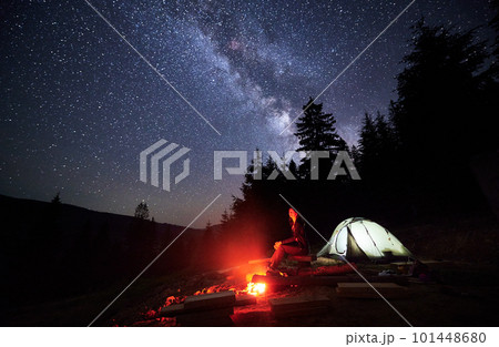 Night-time camping excursion in mountains, amidst twinkling stars. Woman tourist rests by bonfire and tent, nestled near forest, under resplendent canopy of Milky way, beneath mesmerizing night sky. 101448680