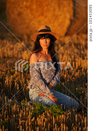A woman with long black hair in jeans, a straw hat sits on her knees in a field A woman with long black hair in jeans, a straw hat sits on her knees in a field 101453095