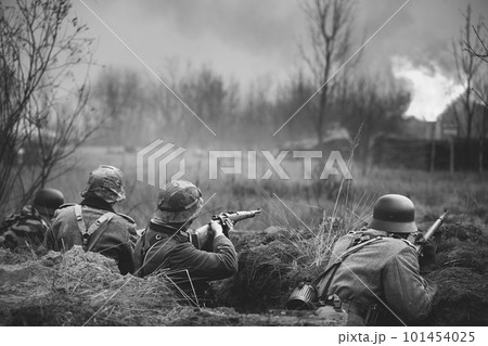 Re-enactors Armed Rifles And Dressed As World War Ii German Wehrmacht Infantry Soldiers Fighting Defensively In Trench. Defensive Position. Building On Fire On Background. Black And White Colors 101454025