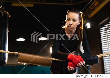 Woman fighter girl power. Woman fighter with red boxing wraps protective bandages standing on boxing ring leaning on ropes waiting resting. Strong powerful girl. Strength fit body workout training 101454267