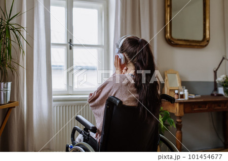 Teenage girl sitting on a wheelchair, listening music and looking out of the window. Teenage girl sitting on a wheelchair, listening music and looking out of the window. 101454677