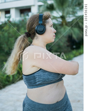Young pretty body positivity plus size woman in sporty top and leggings doing sport fitness and listen to music with headphones in summer morning outdoor 101456191