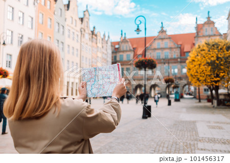 Back view of happy young woman tourist with trendy look with map visiting city and looking around, searching direction on location map during vacation in Gdansk old town. Back view of happy young woman tourist with trendy look with map visiting city and looking around, searching direction on location map during vacation in Gdansk old town. 101456317