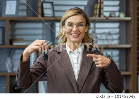Portrait of a senior beautiful businesswoman, realtor, broker, real estate agent standing in the office, holding keys in her hands, pointing at them with her finger, smiling at the camera. Portrait of a senior beautiful businesswoman, realtor, broker, real estate agent standing in the office, holding keys in her hands, pointing at them with her finger, smiling at the camera. 101458048