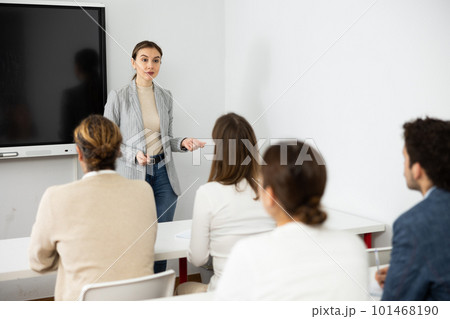 Young female business coach explaining topic while standing against group of people sitting at desks in front of her 101468190