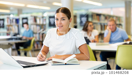 Focused European casual female writing in notepad and using laptop in the library Focused European casual female writing in notepad and using laptop in the library 101468208