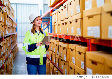Pretty young warehouse worker woman hold tablet and look to product boxes on shelves of stock check 101471880