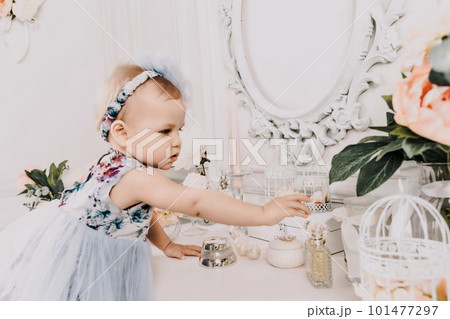 Baby girl elegant dress. A one-year-old girl in a puffy dress and a cute bow poses against the backdrop of a bright room with a dressing table and flowers. 101477297