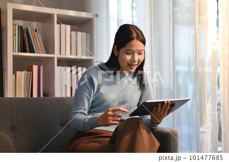 Smiling adult woman sitting on couch browsing wireless internet on digital tablet. Technology, people and lifestyle concept Smiling adult woman sitting on couch browsing wireless internet on digital tablet. Technology, people and lifestyle concept 101477685