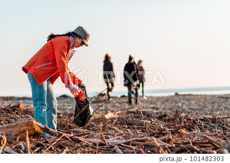A Caucasian woman in a jacket with a garbage bag in her hands is cleaning up on the beach after a storm. In the background, the sea, the sky, and other cleaners. Concept of environmental pollution 101482303