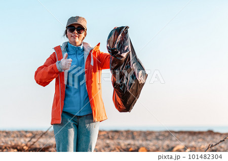 A young Caucasian woman in a jacket, holding a garbage bag and pointing at it with her finger. In the background, the beach after the storm, the sea and the sky 101482304