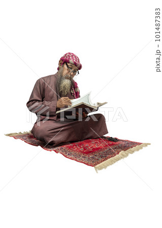 Muslim man with a beard wearing keffiyeh with agal sitting and reading the Quran on the prayer rug Muslim man with a beard wearing keffiyeh with agal sitting and reading the Quran on the prayer rug 101487383