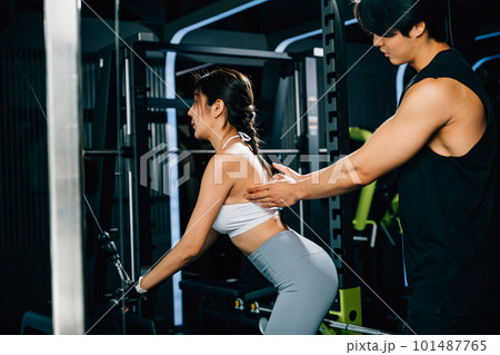 Smiling coach instructing a woman on how to use a pull-down weight machine, focusing on building muscular strength and endurance. GYM healthy lifestyle concept 101487765