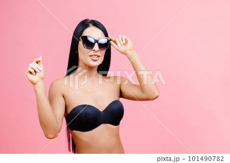 Young woman in summer black swimsuit and sunglasses standing on pink background in studio 101490782