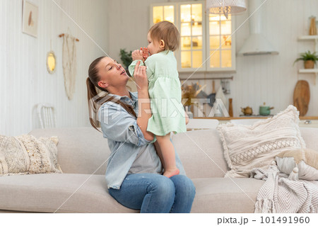 Happy family at home. Mother lifting in air little toddler child daughter. Mom and baby girl playing having fun together at home. Mother hugs baby with love care. Young mother kid rest in living room 101491960