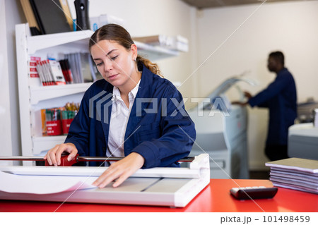 Positive female employee at printing house cuts paper with paper cutter Positive female employee at printing house cuts paper with paper cutter 101498459