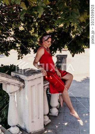 woman in a red silk dress and a bandage on her head smiles against the background of the leaves of a tree. She is leaning on the coop and looking into the camera. Vertical photo. 101509928