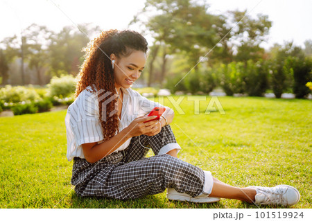 Young woman sitting on green grass in park with phone. Pretty woman listening to music. 101519924