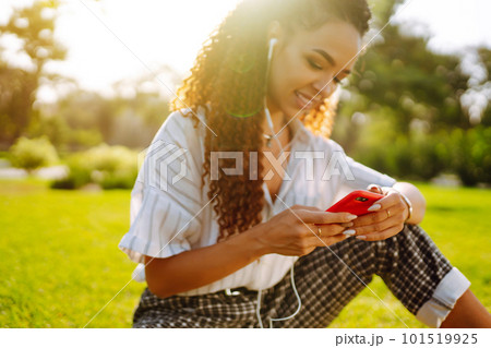 Young woman sitting on green grass in park with phone. Pretty woman listening to music. 101519925