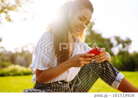 Young woman sitting on green grass in park with phone. Pretty woman listening to music. 101519926
