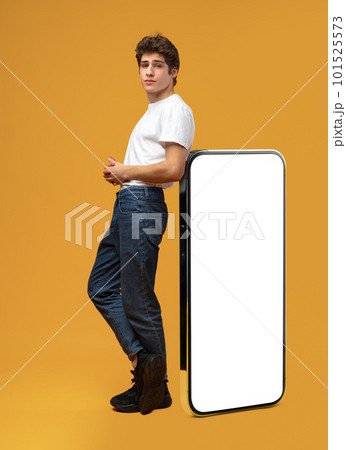 Handsome young man standing leaned on huge smartphone over yellow background Handsome young man standing leaned on huge smartphone over yellow background 101525573