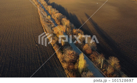 Beautiful landscape view old asphalt road with trees and shadows between large plowed agricultural fields of black soil on sunny autumn evening. Flying over dug agrarian fields of earth. Aerial drone 101528697