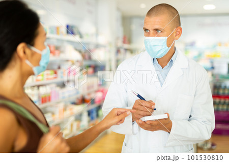 Pharmacist in protective mask writes in notebook the name of the drugs that buyer tells him to 101530810