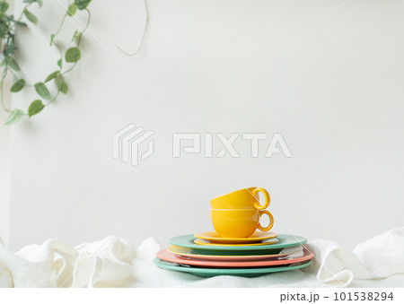 Empty yellow cups and brightly colored plates on a white table covered with a linen tablecloth, a flower pot on the wall in the background. Preparation for serving. 101538294