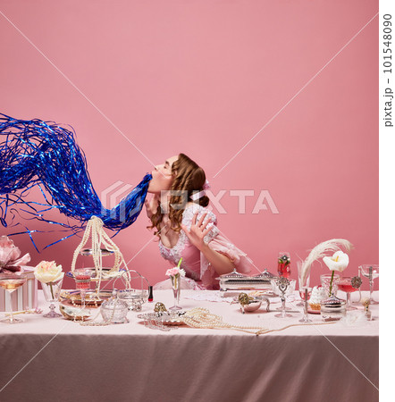 Medieval attractive blond queen wearing fancy pink dress sitting at laid table with hangover over studio background. Alcohol party 101548090