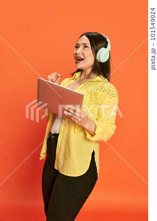Portrait of young emotional girl in headphones and tablet, posing with winning look against orange studio background. Online games Portrait of young emotional girl in headphones and tablet, posing with winning look against orange studio background. Online games 101549024