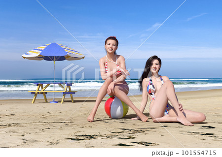 青い空と海を背に大きなパラソルが立つ砂浜で横たわる二人の女の子 101554715