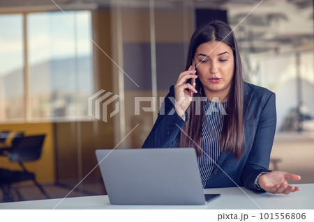 Serious businesswoman talking on mobile phone at office workplace, working at computer. Secretary answering telephone call. Serious businesswoman talking on mobile phone at office workplace, working at computer. Secretary answering telephone call. 101556806