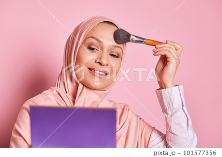 Closeup portrait of a beautiful Middle Eastern Muslim woman in pink hijab, holding palette and using a paintbrush applying makeup foundation and powder on her face, over isolated pink background 101577356