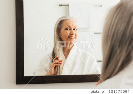 Senior woman examine her long grey hair while standing in front of a bathroom mirror	 101579094