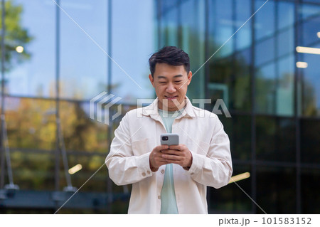 Portrait of young Asian man designer, freelancer, businessman standing near office center and using phone, reading news, writing message, checking mail, waiting for meeting. Portrait of young Asian man designer, freelancer, businessman standing near office center and using phone, reading news, writing message, checking mail, waiting for meeting. 101583152