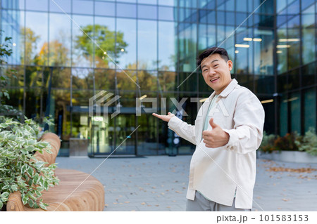 A young Asian man, a businessman, a freelancer, is standing outside an office, a skyscraper, and points to the entrance with his hands. invites with a gesture to come inside, see his project, work. A young Asian man, a businessman, a freelancer, is standing outside an office, a skyscraper, and points to the entrance with his hands. invites with a gesture to come inside, see his project, work. 101583153